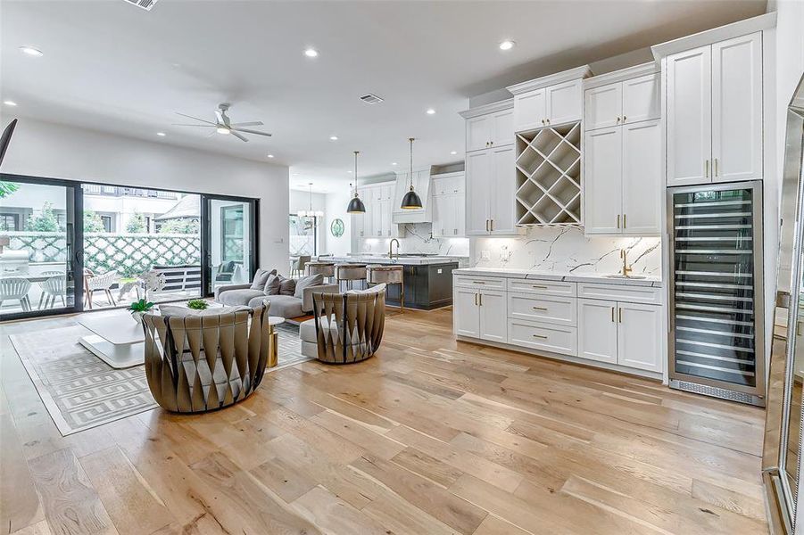 This modern open-concept kitchen and living area features two Cantori twist chairs, sleek white custom cabinetry, a wet bar, a large quartz kitchen island, and a wine fridge. The space is brightened by natural light from expansive sliding glass doors leading to a spacious outdoor patio. The light wood flooring and contemporary design elements create an inviting atmosphere.