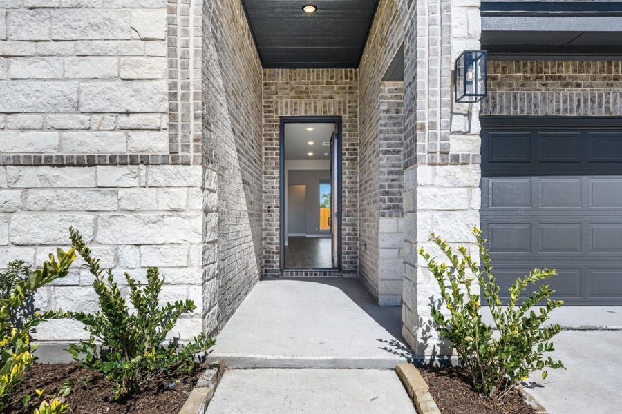 Covered front entry with brick and stone exterior, open door, wall lantern, and dark garage door nearby Covered front entry with brick and stone exterior, open door, wall lantern, and dark garage door nearby
