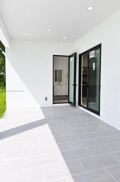 Covered patio featuring large-format grey tile flooring, recessed ceiling lighting, black-framed glass sliding doors, and a full-height glass-panel swing door