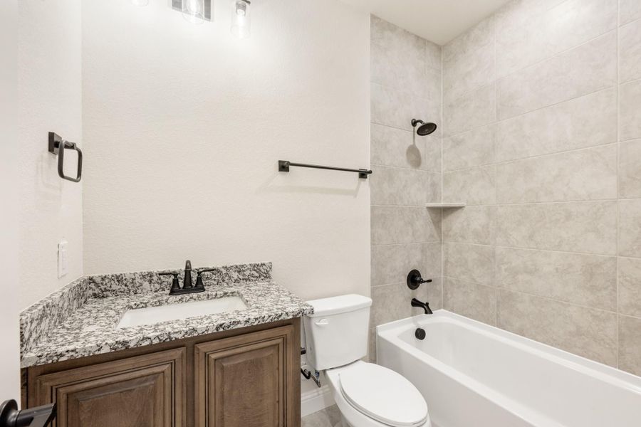 Bathroom with granite vanity, wood cabinets, toilet, and tiled shower-tub combo with dark fixtures