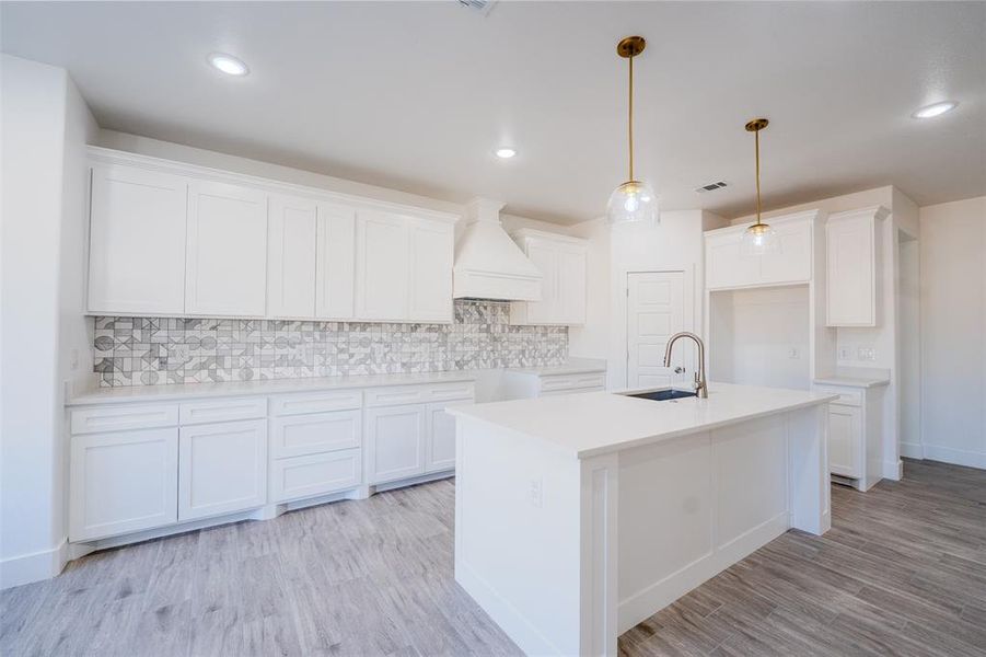 Kitchen with tasteful backsplash, pendant lighting, custom exhaust hood, white cabinetry, and an island with sink Kitchen with tasteful backsplash, pendant lighting, custom exhaust hood, white cabinetry, and an island with sink