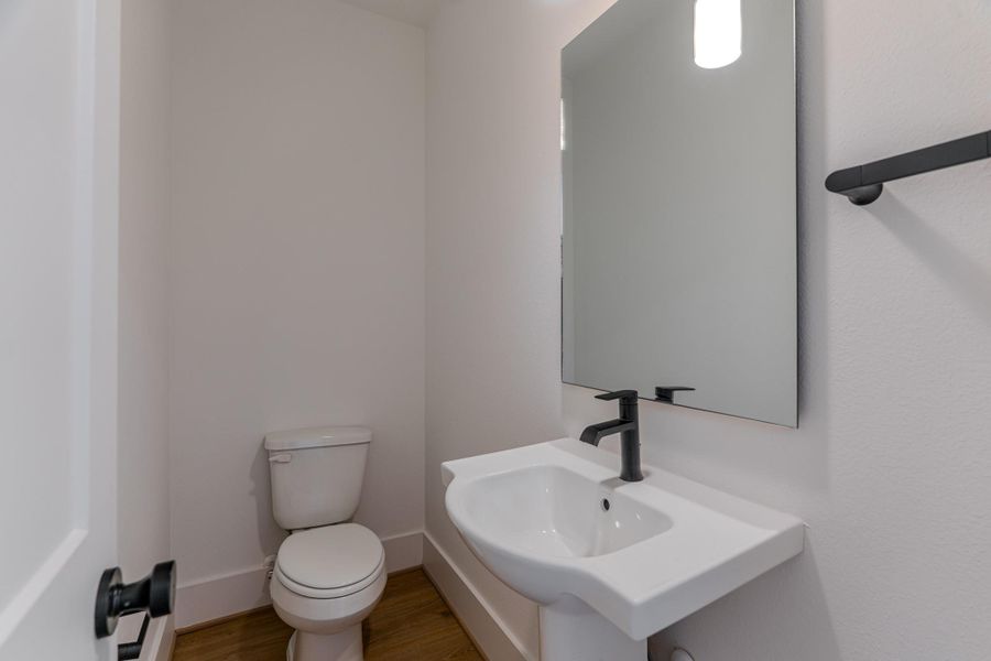 This photo shows a compact, modern half-bathroom with a toilet and a pedestal sink. It features a sleek black faucet, a large mirror, and light-colored walls, creating a clean and contemporary look. The space has wooden flooring, adding warmth to the minimal design.Photo Disclaimer: Photos are representative of the community and may depict a similar floor plan, model home, or previously completed residence. This photo shows a compact, modern half-bathroom with a toilet and a pedestal sink. It features a sleek black faucet, a large mirror, and light-colored walls, creating a clean and contemporary look. The space has wooden flooring, adding warmth to the minimal design.Photo Disclaimer: Photos are representative of the community and may depict a similar floor plan, model home, or previously completed residence.