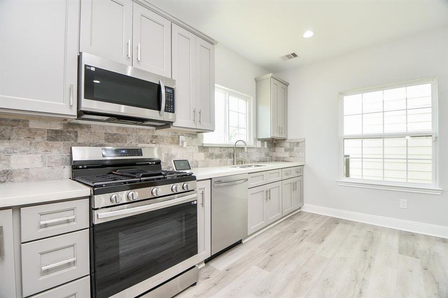 This is a modern, bright kitchen with white cabinetry, stainless steel appliances, a gas range, and a stylish stone backsplash. The room features light wood flooring and a large window that lets in plenty of natural light.