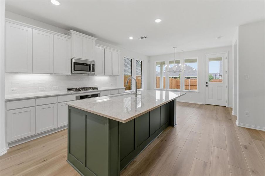 Contemporary kitchen featuring a large central island with a contrasting dark green base, white shaker-style cabinetry, stainless steel appliances, light-colored countertops, and wood-finish flooring