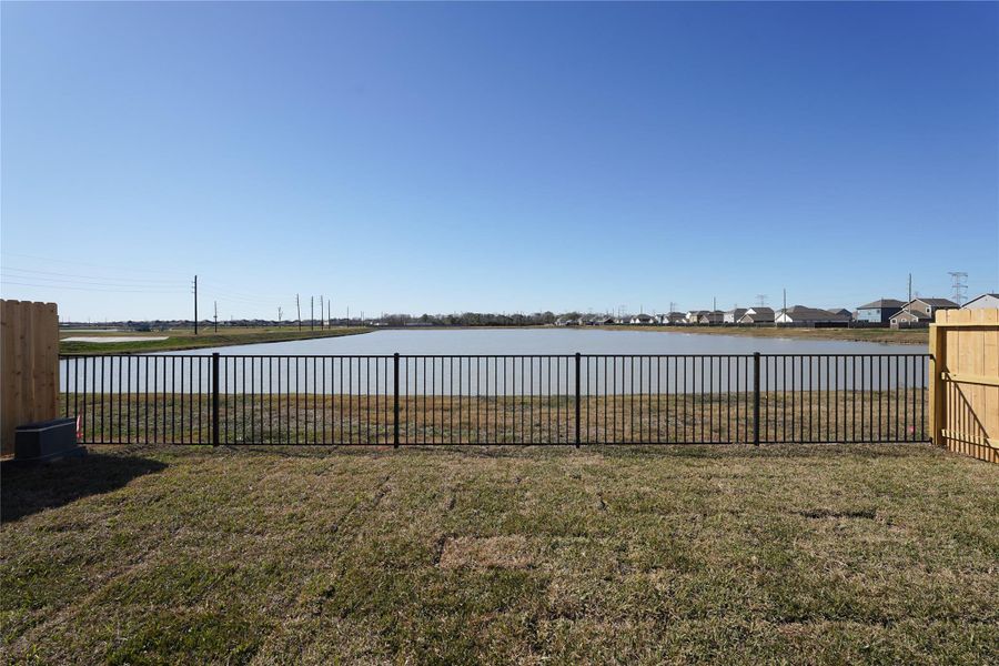 The photo showcases a backyard with a metal fence overlooking a serene pond, offering a peaceful view and a spacious grassy area perfect for outdoor activities.