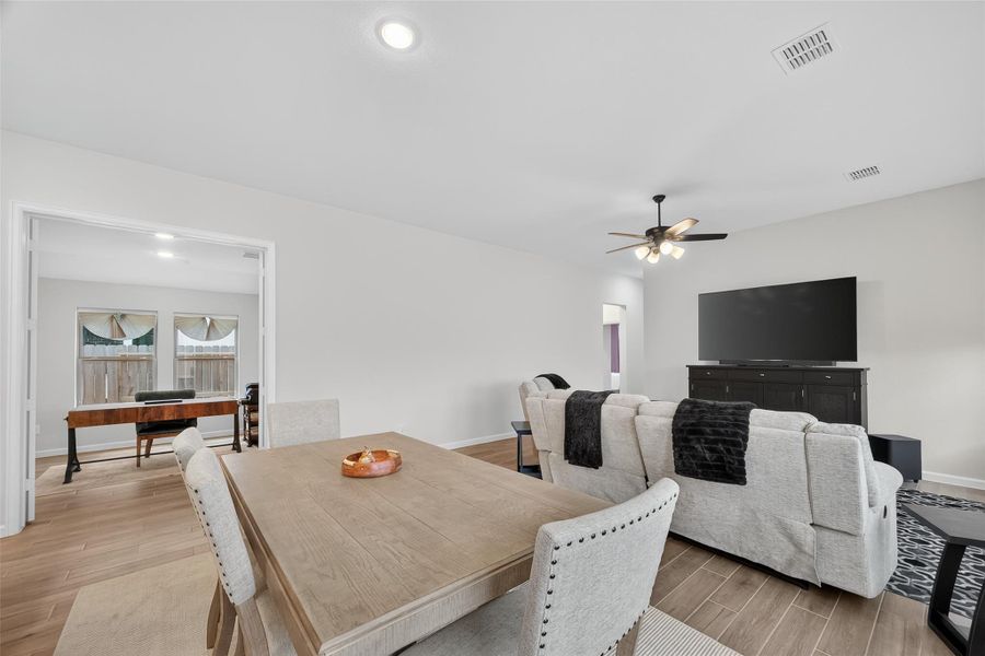 This spacious living area features an open layout with light wood like flooring, a cozy seating area with a large TV, and a dining space. A ceiling fan provides comfort, while double doors lead to a well-lit room, perfect for an office or playroom. This spacious living area features an open layout with light wood like flooring, a cozy seating area with a large TV, and a dining space. A ceiling fan provides comfort, while double doors lead to a well-lit room, perfect for an office or playroom.