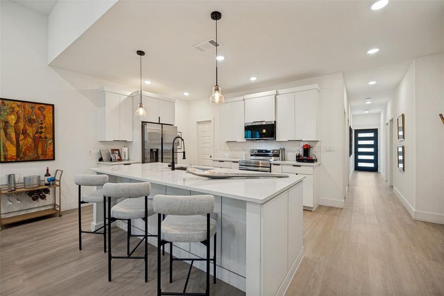 Modern kitchen featuring a large central island with seating, white shaker-style cabinetry, stainless steel appliances, and sleek pendant lighting Modern kitchen featuring a large central island with seating, white shaker-style cabinetry, stainless steel appliances, and sleek pendant lighting