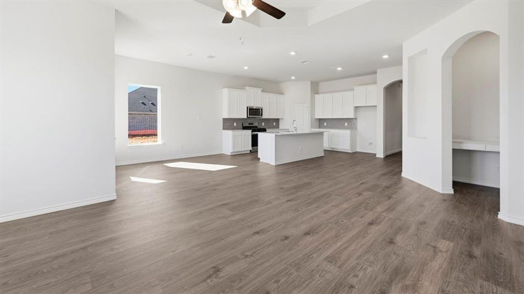 Spacious open-concept interior featuring wood-finish flooring, a contemporary kitchen with a central island and white cabinetry, a built-in desk niche, recessed lighting, and a ceiling fan