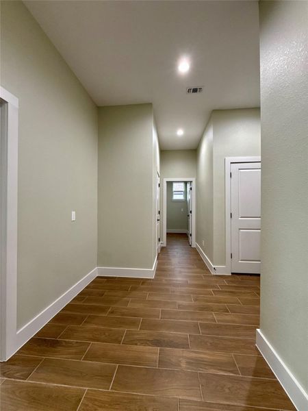 A spacious hallway features wood-look tile flooring and neutral-colored walls. It is well-lit with recessed lighting and has several doorways leading to other rooms.