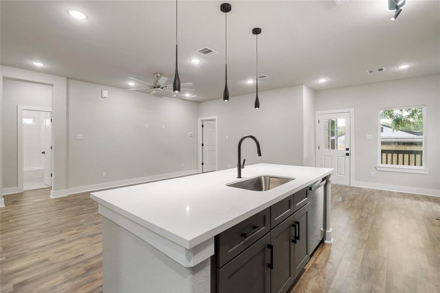 This modern, open-concept living space features a sleek kitchen island with a sink, pendant lighting, and wood-style flooring. The area is bright with natural light, offering a seamless flow to an adjoining room and access to the backyard through a glass door. This modern, open-concept living space features a sleek kitchen island with a sink, pendant lighting, and wood-style flooring. The area is bright with natural light, offering a seamless flow to an adjoining room and access to the backyard through a glass door.