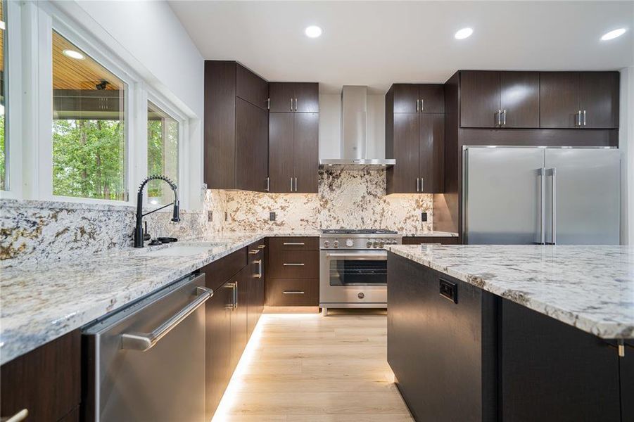 Kitchen featuring high end appliances, wall chimney range hood, light wood-type flooring, decorative backsplash, and light stone counters Kitchen featuring high end appliances, wall chimney range hood, light wood-type flooring, decorative backsplash, and light stone counters