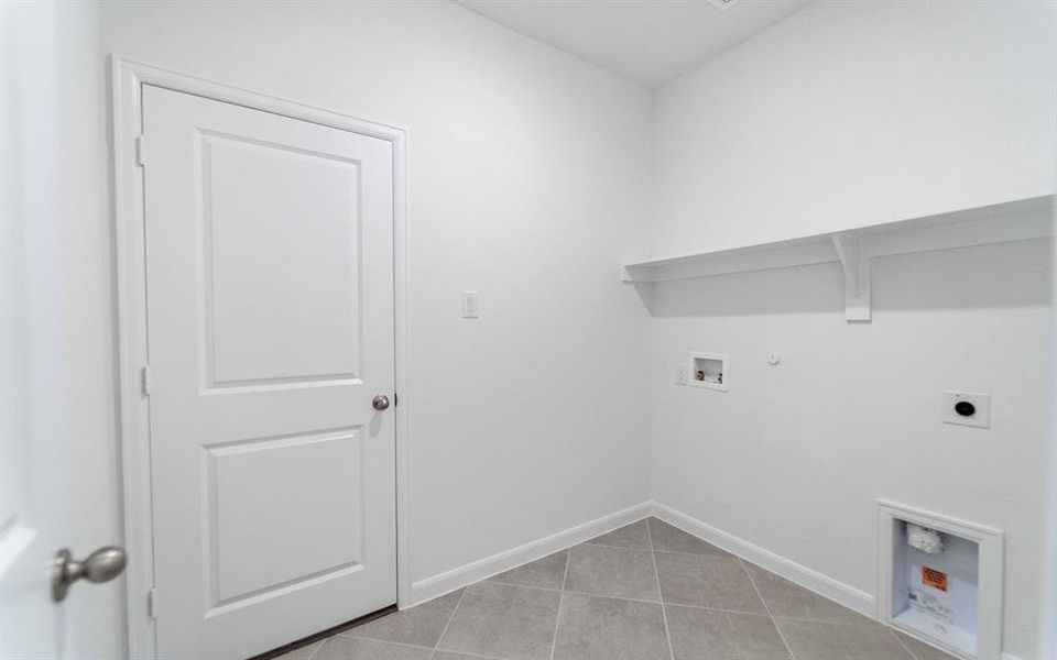 Utility area featuring an overhead storage shelf, tiled flooring, white walls, and a white door with a brushed nickel knob