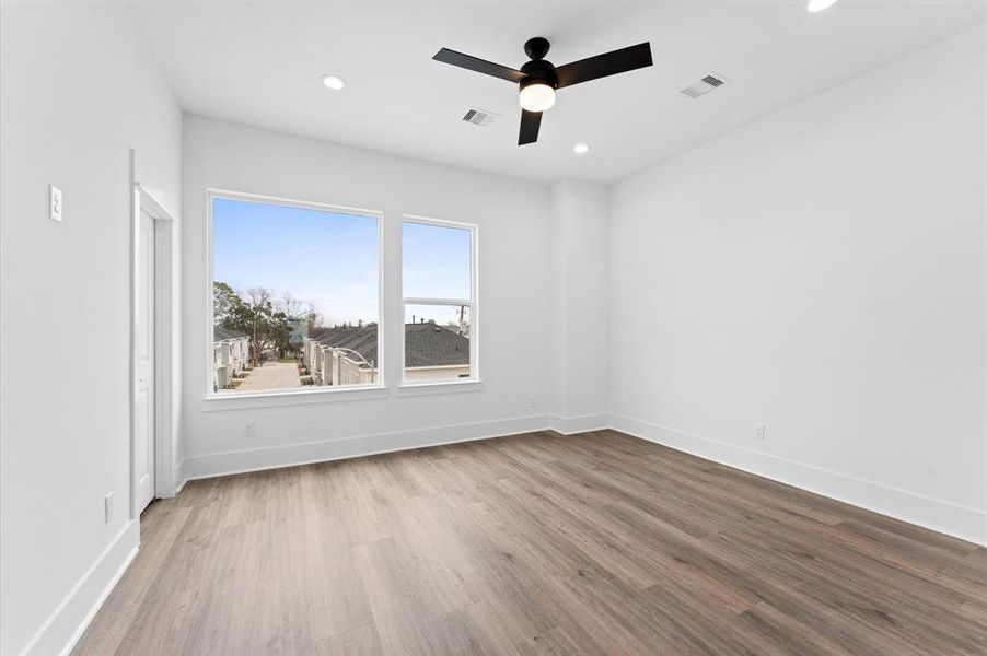 This is a bright, spacious room featuring large windows with a view, light wood flooring, and a modern ceiling fan. The walls are painted white, enhancing the natural light.