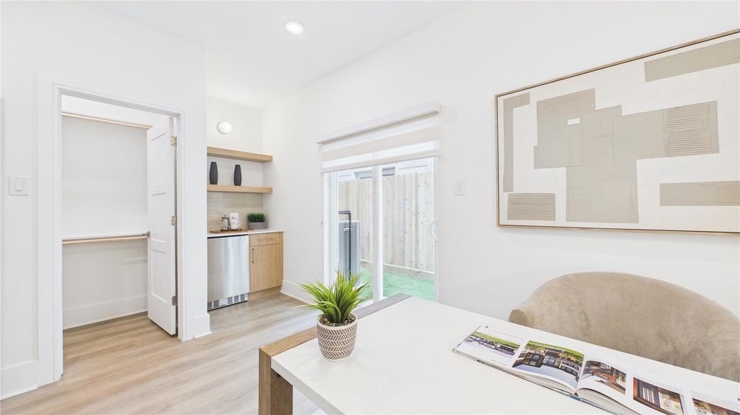 Bright and modern room with light wood flooring, featuring a cozy seating area and a sleek kitchenette. The space includes a large window with a view of a fenced outdoor area, and minimalist decor enhances the open feel.