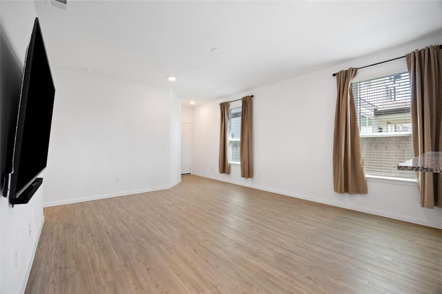 Spacious living area with light wood flooring, large windows with brown curtains, and a wall-mounted TV, offering a bright and inviting atmosphere.