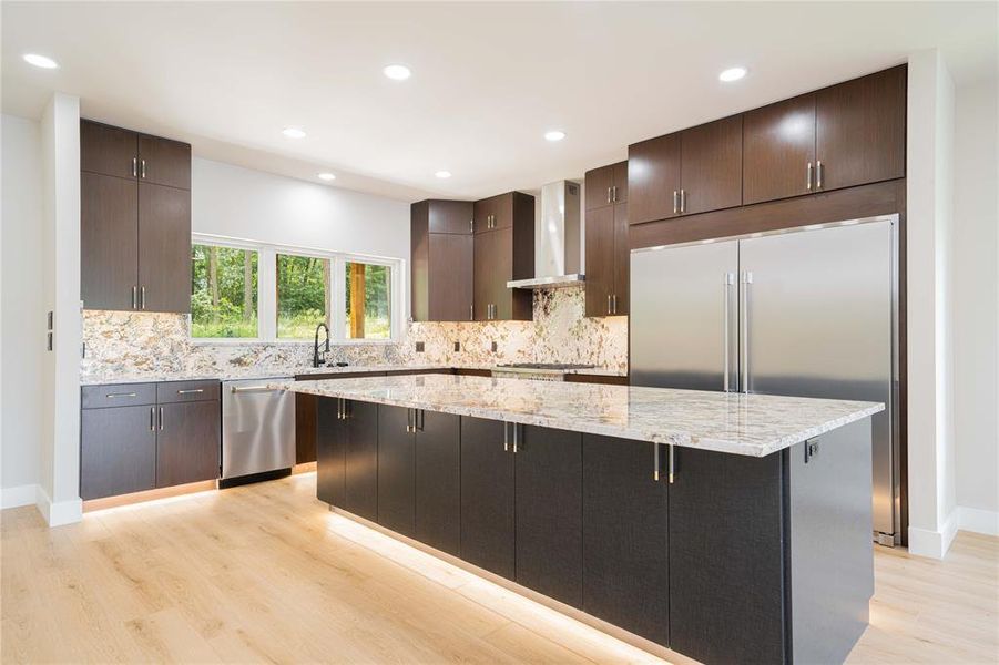 Kitchen with wall chimney exhaust hood, appliances with stainless steel finishes, light wood-style floors, a kitchen island, and dark brown cabinets Kitchen with wall chimney exhaust hood, appliances with stainless steel finishes, light wood-style floors, a kitchen island, and dark brown cabinets