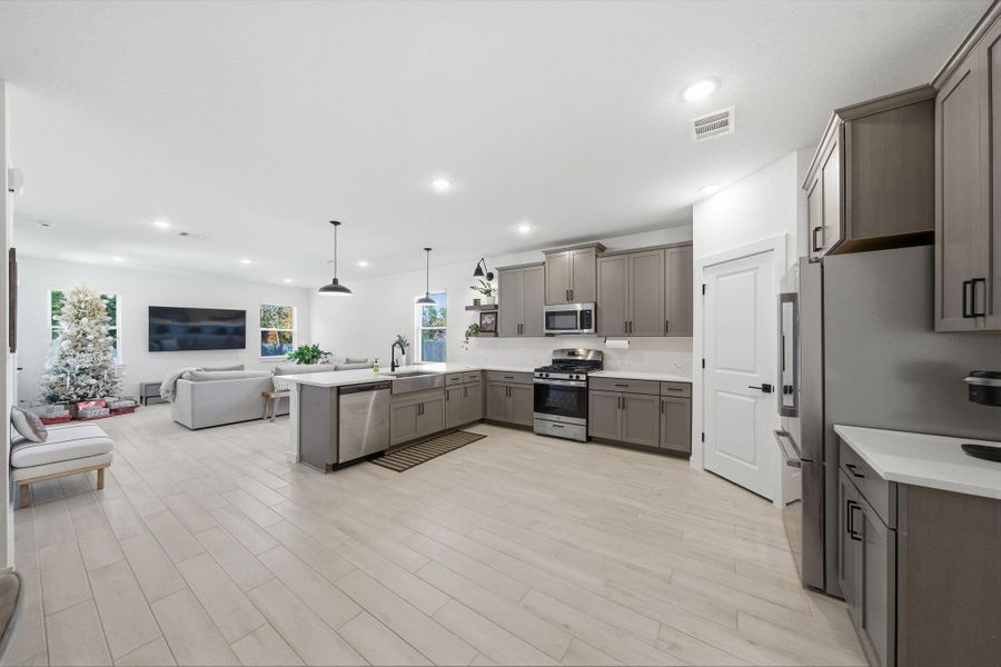 This photo showcases a spacious, open-concept kitchen and living area. The kitchen features modern gray cabinets, stainless steel appliances, and an island with pendant lighting. The adjacent living area has large windows, a wall-mounted TV, and a decorated Christmas tree, providing a cozy and inviting atmosphere.