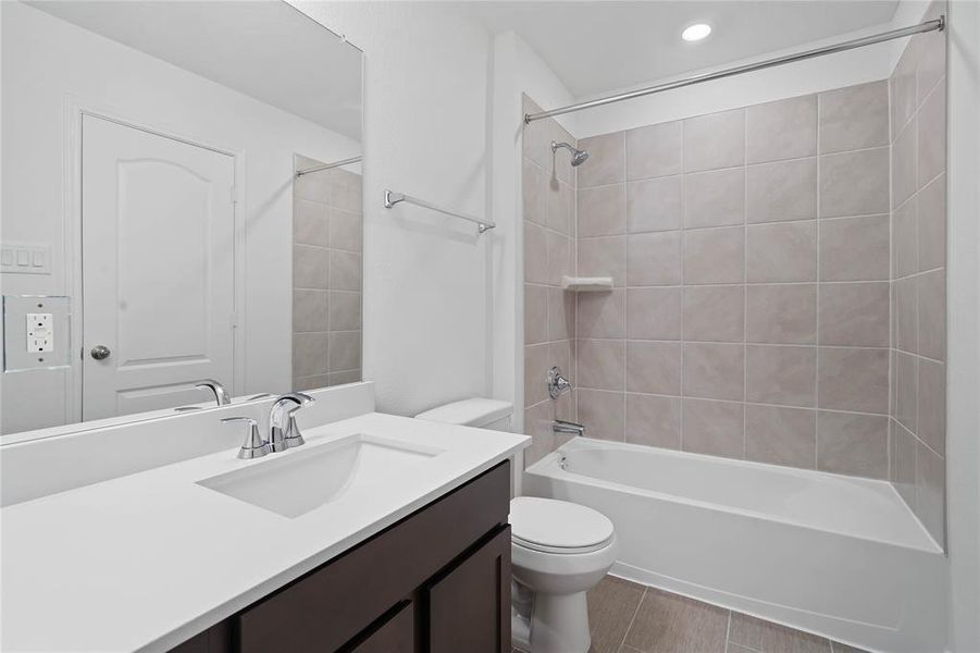 Secondary bath features tile flooring, bath/shower combo with tile surround, stained wood cabinets, beautiful light countertops, mirror, dark, sleek fixtures and modern finishes.