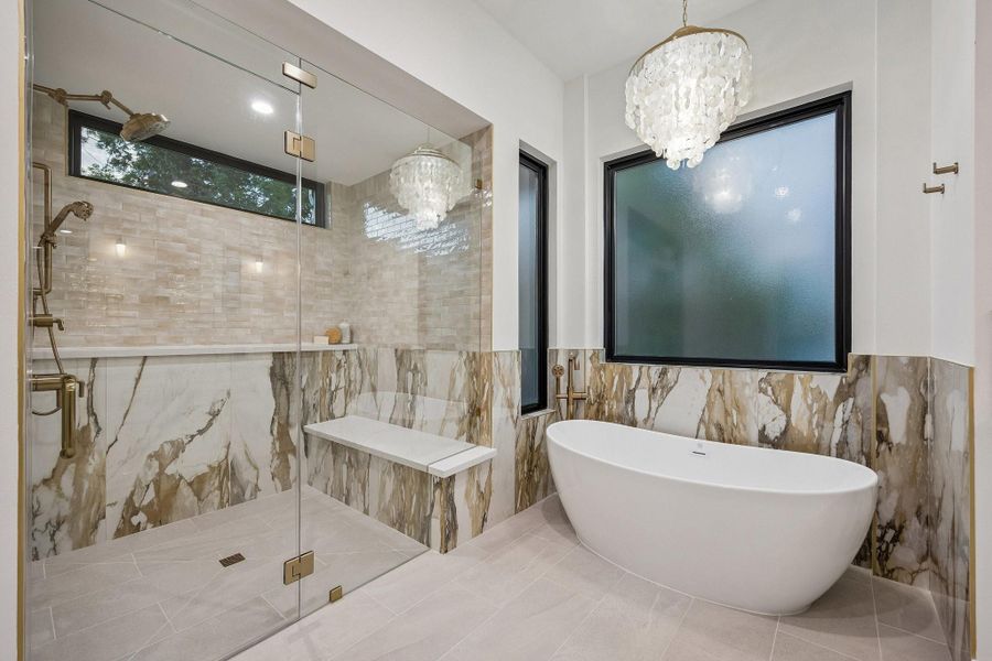 Large Walk-in Shower with a Frameless Shower Enclosure. The 12 x 24 Matt Tile Bathroom Floor continues into the Shower. The Shower walls are a mixture 2 x 6 Glossy Tile and 24 x 48 Macchia Vecchia Polished Tile. The Macchia Vecchia Polished 24 x 48 Tile continues around the Free-Standing Soaking Tub with a Floor-Mounted Tub Filler in Luxe Gold. Above the tub hangs a stunning tiered 24" Gold Coastal Capiz Natural Seashell Chandelier.