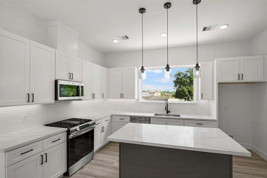 This modern kitchen features sleek white cabinetry, stainless steel appliances, and a spacious island with pendant lighting. It offers ample natural light through a large window, creating a bright and inviting space.