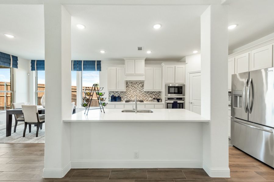 Kitchen with white cabinets, herringbone tile backsplash, stainless steel appliances, and white island with sink open to dining area.