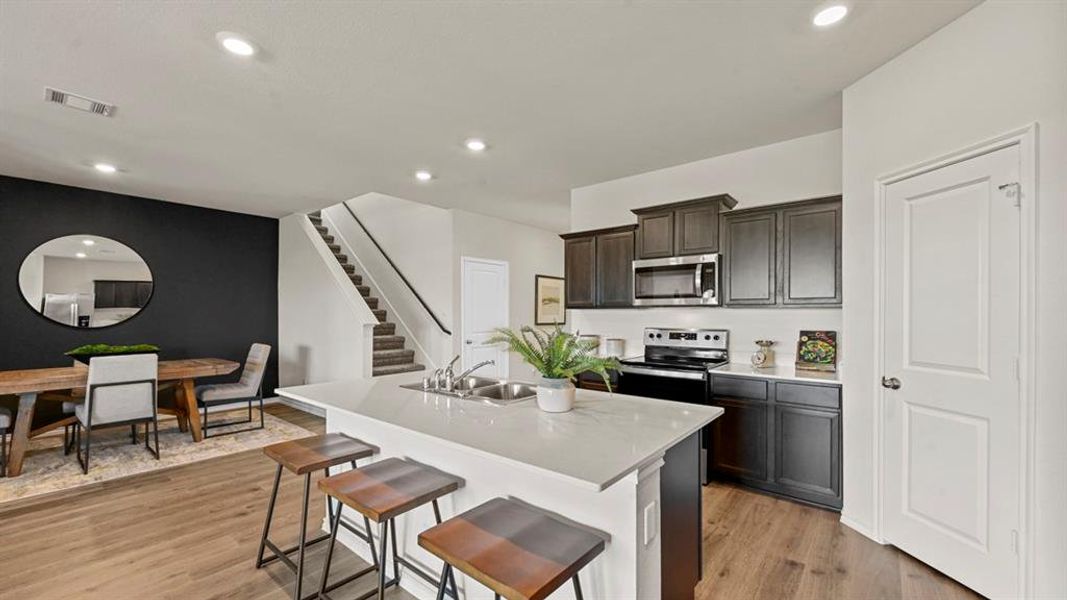 The open-concept living area features wood-look flooring, a kitchen with dark cabinetry and stainless steel appliances, and a dining area with a contrasting dark accent wall The open-concept living area features wood-look flooring, a kitchen with dark cabinetry and stainless steel appliances, and a dining area with a contrasting dark accent wall