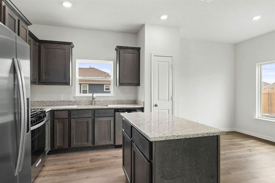 Kitchen featuring appliances with stainless steel finishes, light stone countertops, light wood finished floors, dark brown cabinetry, and a center island