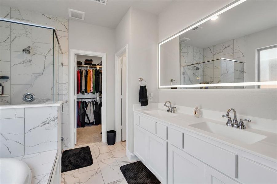 Bathroom featuring a double vanity with white cabinetry, a large illuminated mirror, and a walk-in shower with glass enclosure