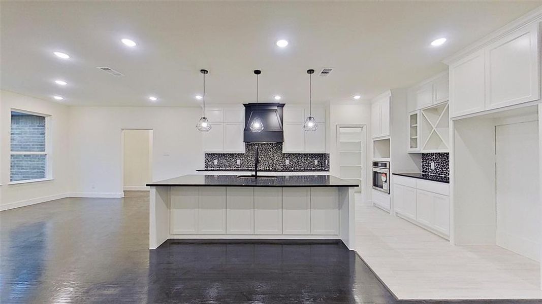 Kitchen with a large island with sink, tasteful backsplash, white cabinets, hanging light fixtures, and stainless steel oven Kitchen with a large island with sink, tasteful backsplash, white cabinets, hanging light fixtures, and stainless steel oven