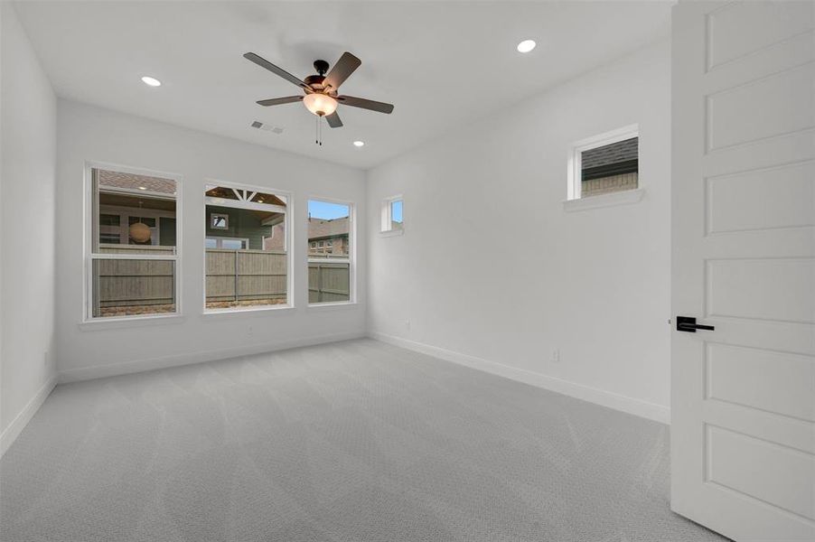 Spacious room featuring light gray carpet, white walls, multiple windows providing natural light, and a ceiling fan with integrated lighting