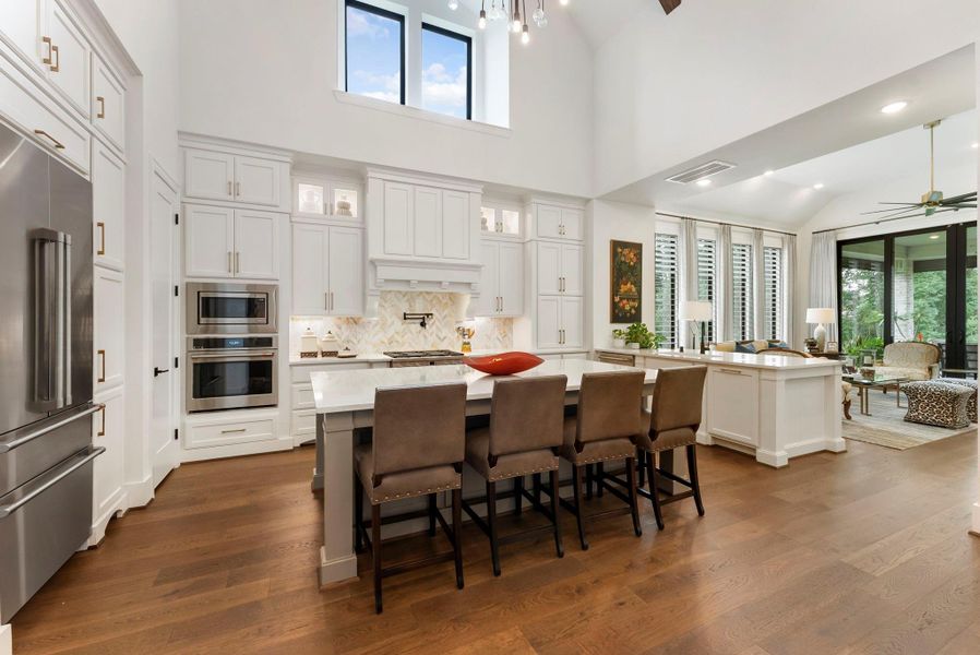 Soaring ceilings, exposed beams, and an open kitchen design create a dramatic central gathering space. The kitchen features upgraded cabinetry, a large island, built-in refrigerator, and marble backsplash for a timeless yet current feel.