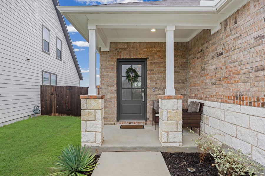 Covered front porch featuring a dark gray paneled door with glass inserts, brick and stone veneer siding, white columns, and concrete flooring