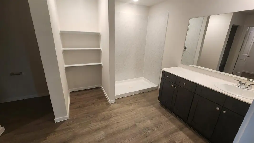 Spacious bathroom featuring wood-finish flooring, a contemporary dark wood vanity with a white countertop and integrated sink, a large wall-mounted mirror, a tiled shower enclosure with a white base, and built-in shelving