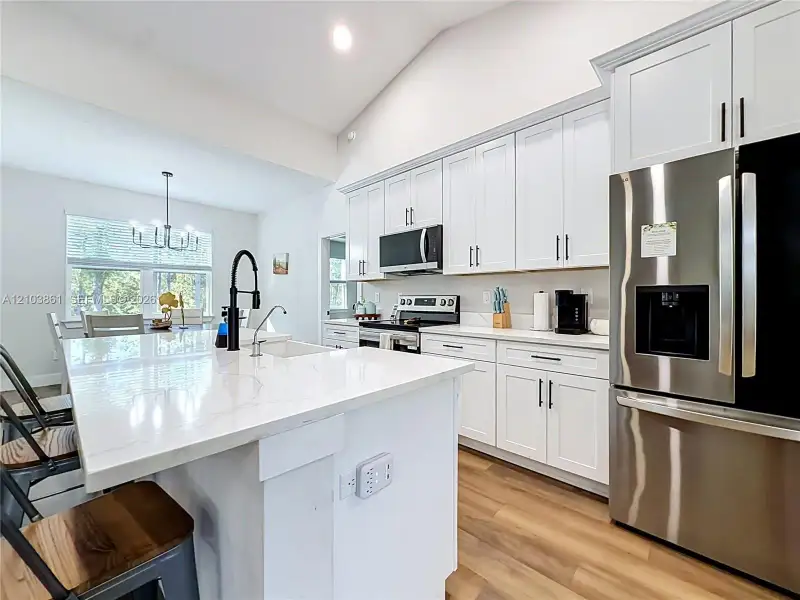 Kitchen featuring white Shaker-style cabinetry, a central island with a solid-surface countertop, stainless steel appliances, a matte black high-arc faucet, and wood-finish flooring