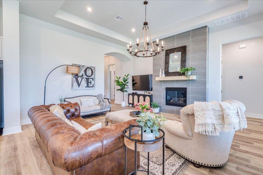 Living room featuring a tray ceiling, a tiled fireplace, light wood-style flooring, suspended lighting, and arched walkways Living room featuring a tray ceiling, a tiled fireplace, light wood-style flooring, suspended lighting, and arched walkways