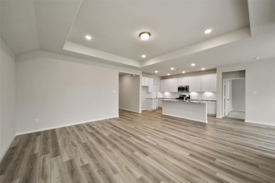 This photo showcases a spacious, open-concept living area with modern wood flooring and a bright kitchen featuring white cabinetry, granite countertops, and stainless steel appliances. The recessed lighting and neutral walls add a touch of elegance, creating a welcoming and versatile space.