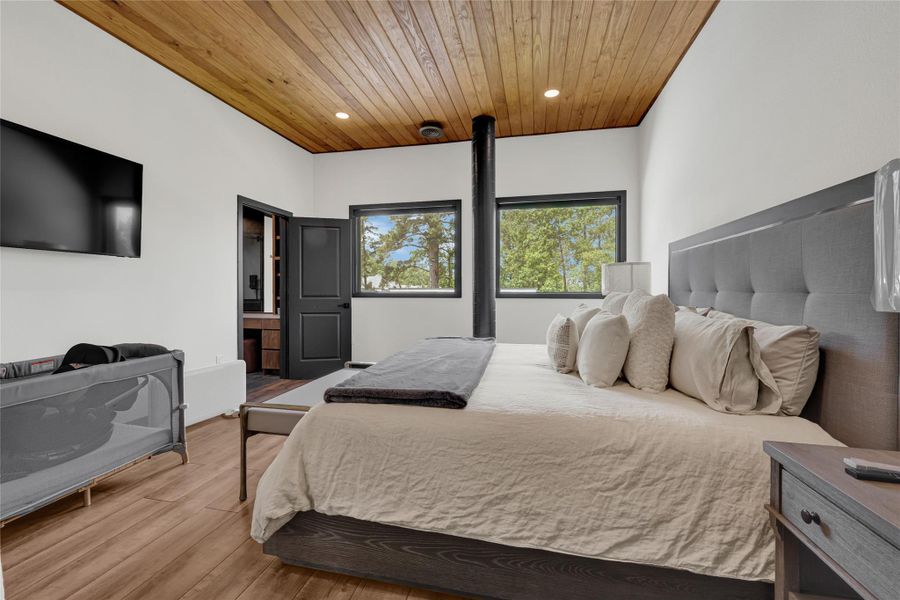 Spacious bedroom with a modern, cozy feel. Features a wooden ceiling, large windows with scenic views, a sleek black chimney, and a mounted TV. Natural light complements the neutral tones and comfortable bedding.