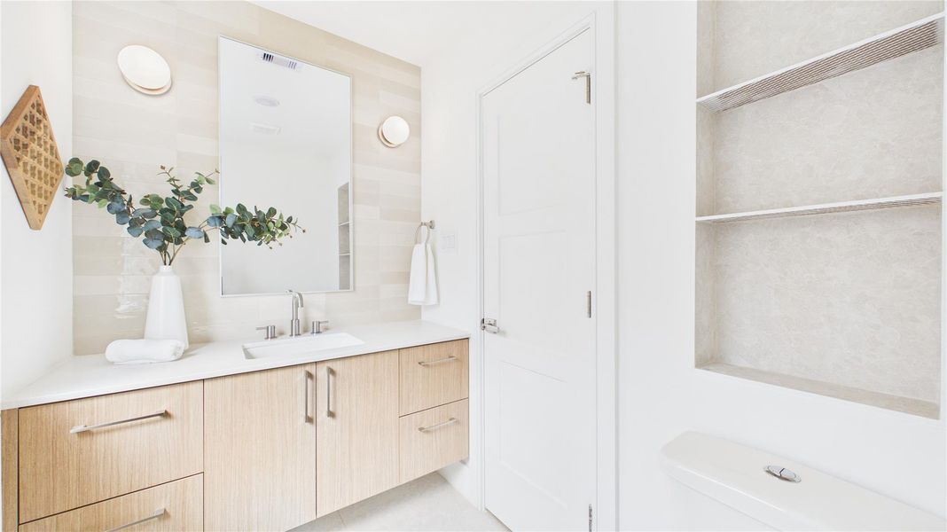 This bathroom features a modern design with a light wood vanity, sleek countertop, and integrated sink. It includes a large mirror, wall-mounted lights, open shelving, and a contemporary toilet. The bright, neutral tones create a clean, airy feel.