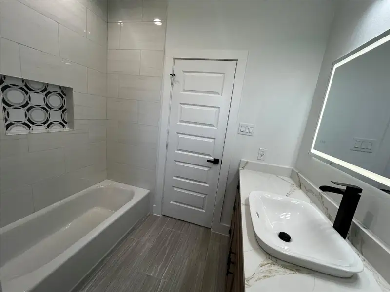 Bright and modern bathroom featuring a sleek bathtub with a decorative tile niche, a stylish vanity with a vessel sink, and an illuminated mirror. The space is enhanced by light tile flooring and contemporary fixtures.