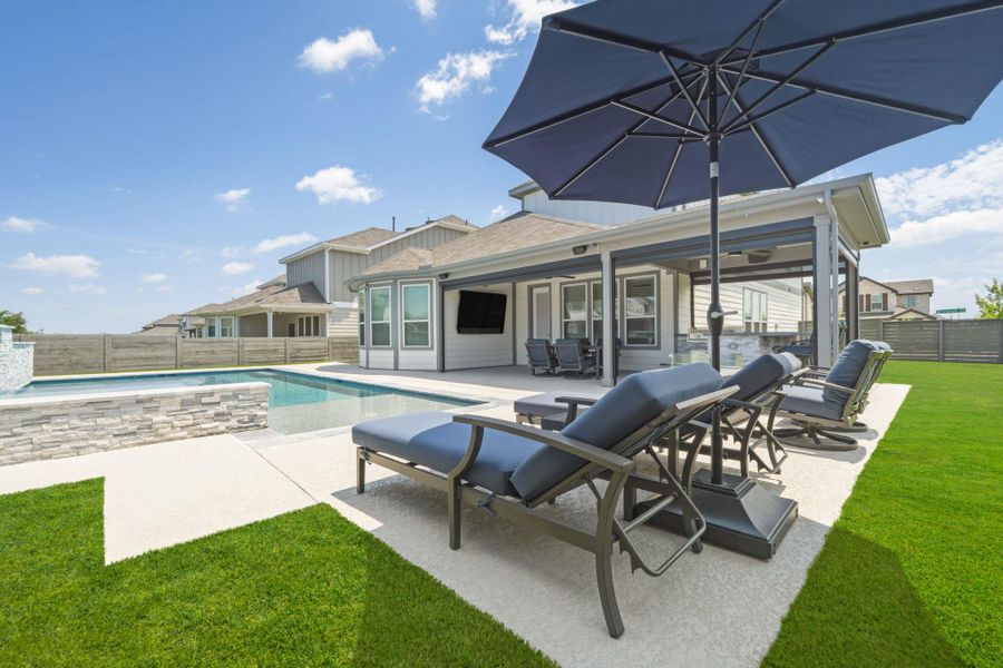 Outdoor pool area featuring a rectangular pool with stone-clad surround, spacious concrete patio, covered outdoor living space, and an expansive turf lawn