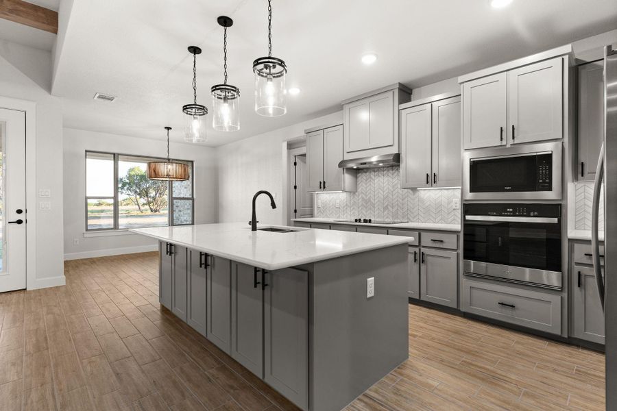 Kitchen with gray cabinets, backsplash, appliances with stainless steel finishes, a center island with sink, and hanging light fixtures Kitchen with gray cabinets, backsplash, appliances with stainless steel finishes, a center island with sink, and hanging light fixtures