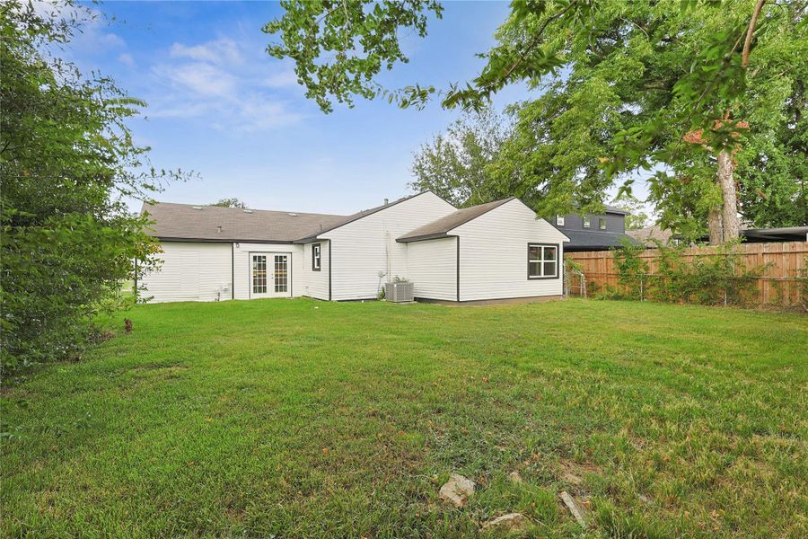 Spacious backyard with lush grass, surrounded by a wooden fence and mature trees. The back of the house features white siding and a set of French doors, providing lots of natural light. Ideal for outdoor activities and relaxation.