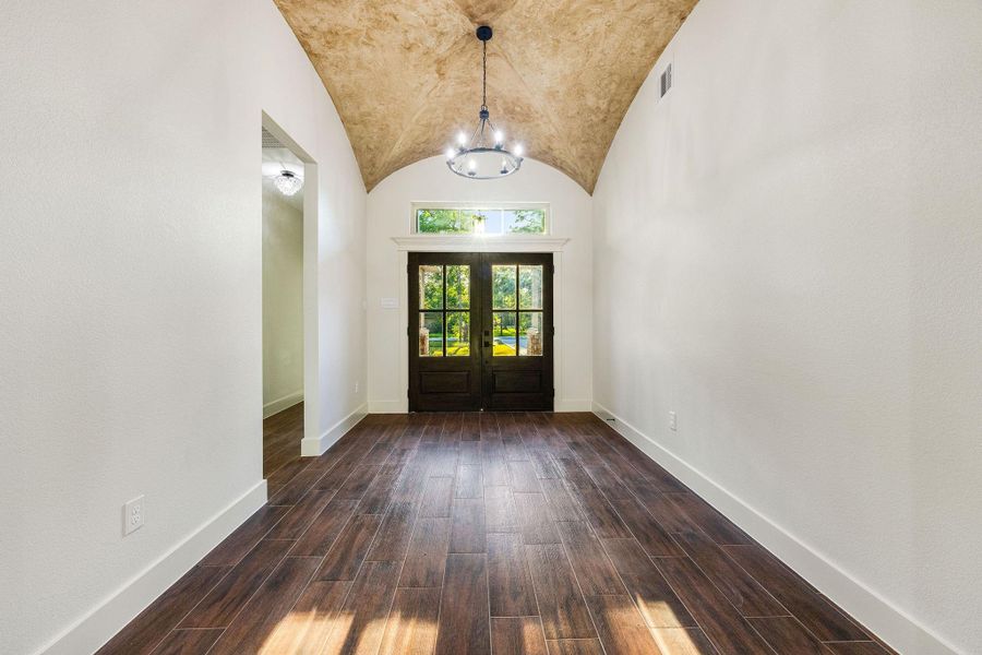 This inviting entryway features a high, textured ceiling and elegant chandelier. The rich wood flooring complements the dark double doors, creating a warm and welcoming ambiance.