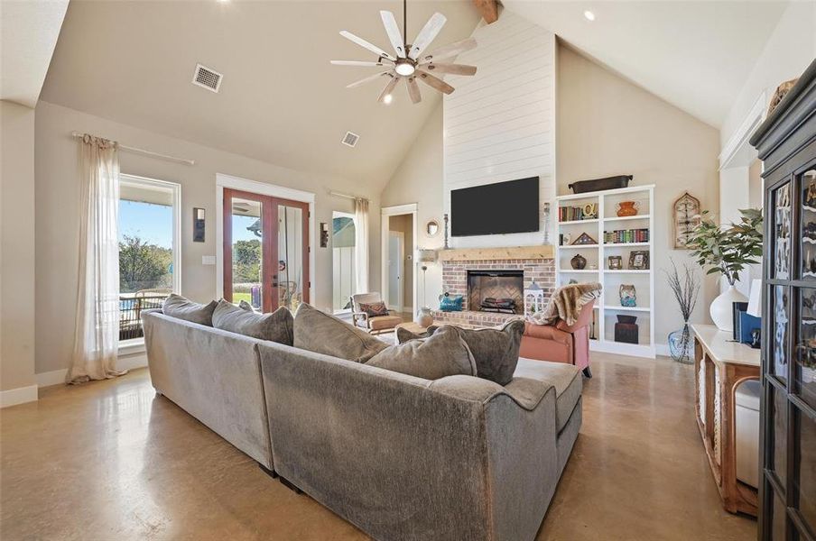 Living room featuring french doors, high vaulted ceiling, a brick fireplace, a ceiling fan, and finished concrete floors Living room featuring french doors, high vaulted ceiling, a brick fireplace, a ceiling fan, and finished concrete floors