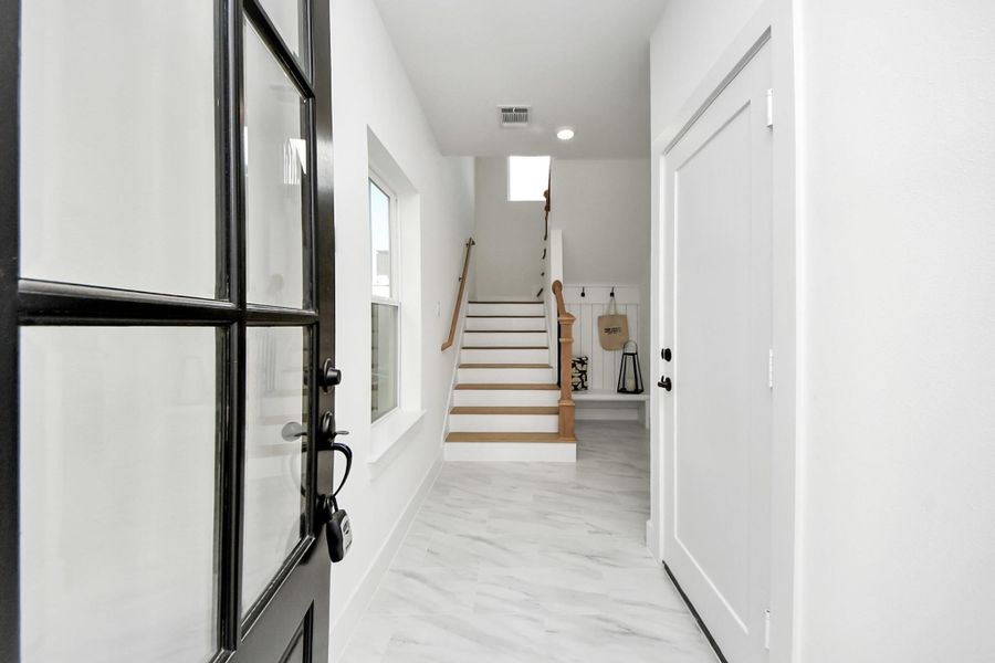 This photo showcases a bright, modern entryway with white walls and marble flooring. A black-framed glass door opens into the space, leading to a staircase with wooden handrails. The area is well-lit and features a window for natural light. This photo showcases a bright, modern entryway with white walls and marble flooring. A black-framed glass door opens into the space, leading to a staircase with wooden handrails. The area is well-lit and features a window for natural light.