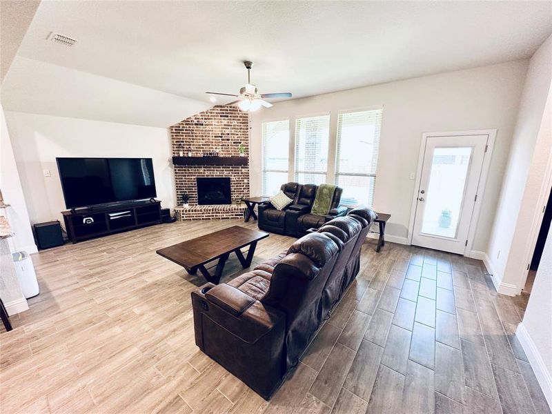 Living room featuring light hardwood / wood-style flooring, ceiling fan, brick wall, a fireplace, and vaulted ceiling