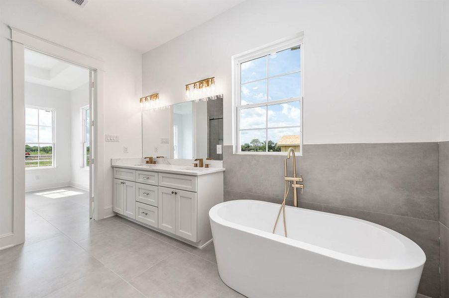 Another view of the primary bathroom featuring a freestanding soaking tub, double vanity, gold fixtures, and a window that brings in natural light.