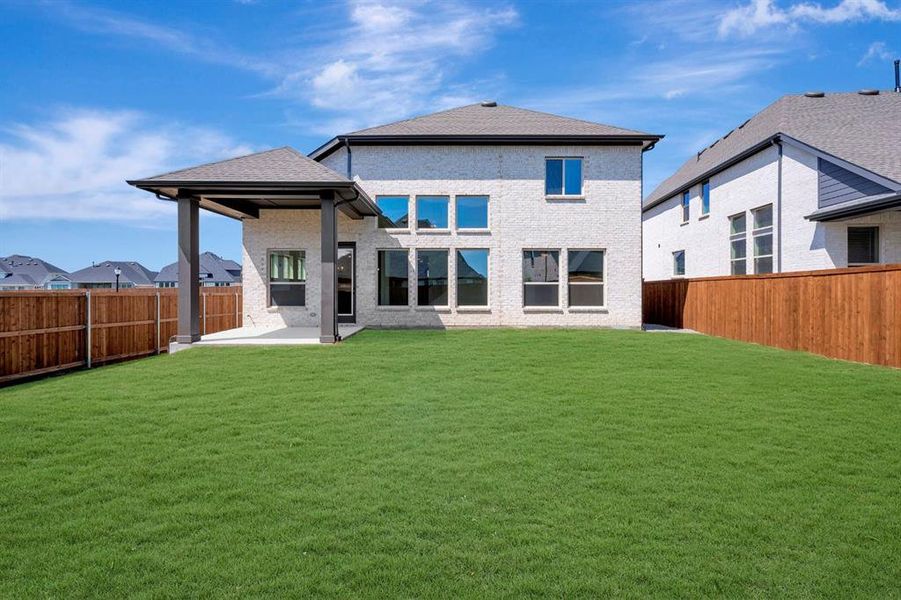 Expansive green lawn with a covered patio, featuring a light-colored brick exterior with dark trim, multiple large windows, and a privacy fence