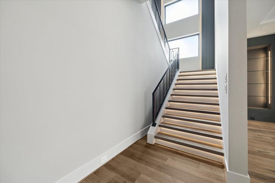 The stairway to the second floor is a stunning architectural element, featuring lighted stair treads that add both safety and dramatic effect. A modern iron railing complements the sleek design, while large glass-pane windows with a lighted accent feature flood the space with natural light and create a striking contemporary focal point.