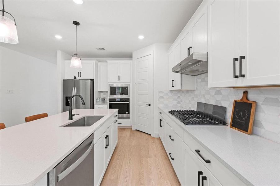 Bright kitchen featuring white cabinetry with dark hardware, a center island with an undermount sink, light wood-finish flooring, and a patterned tile backsplash Bright kitchen featuring white cabinetry with dark hardware, a center island with an undermount sink, light wood-finish flooring, and a patterned tile backsplash